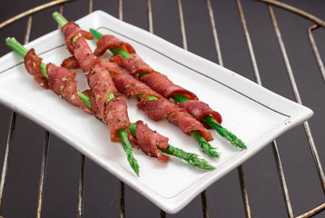 Asparagus wrapped in bacon with spices, grilled on white plate on dark background