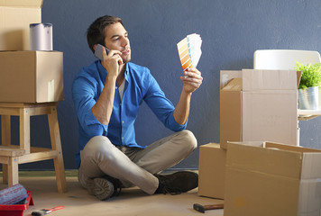Moving young man with mobile phone. Shot of a confident young man with handy sitting in her new home surrounded by cardboard box while matchning the best wall color.