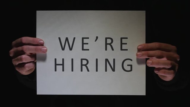 WE'RE HIRING sign being showed to the camera by two hands. Black background