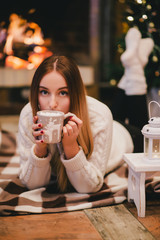 Beautiful young woman sitting near fireplace under the Christmas tree drinking cocoa with marshmallow. Cozy mood