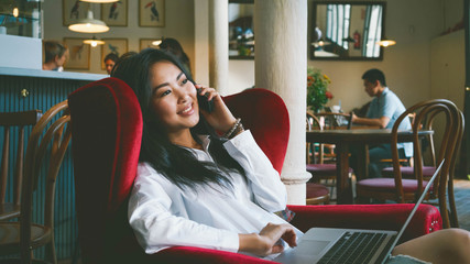 A successful freelancer is sitting in a modern coffee shop with the electronic devices. An asian...