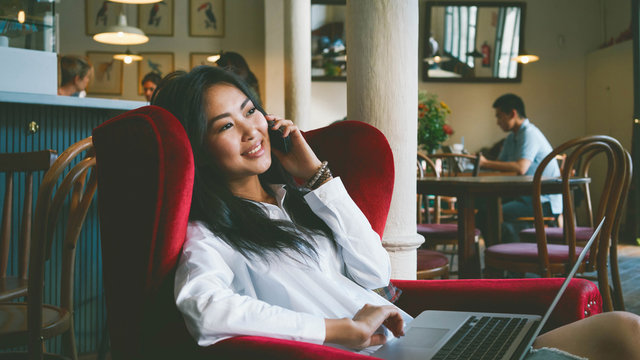 A successful freelancer is sitting in a modern coffee shop with the electronic devices. An asian girl with a laptop is talking by the mobile phone with a friend while having coffee break.