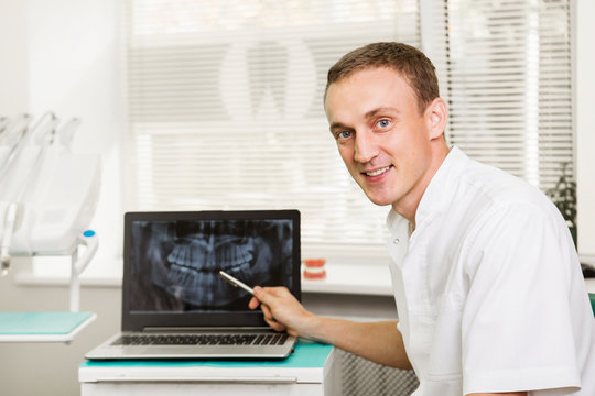  Attractive Male dentist  with perfect smile showing on a laptop teeth x-ray picture in dental clinic office. stomatology and health care concept