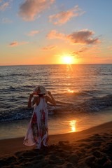 Mädchen im langen  Kleid und hält mit beiden Händen Hut auf dem Kopf und steht am Meeres Strand  beim Sonnenuntergang, mit Blick zur Sonne