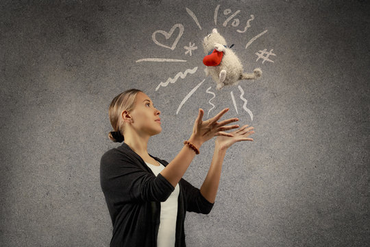 Young beautiful blond woman tosses plush toy cat with plush heart. Doodle sketch is drawn on grey texture background.