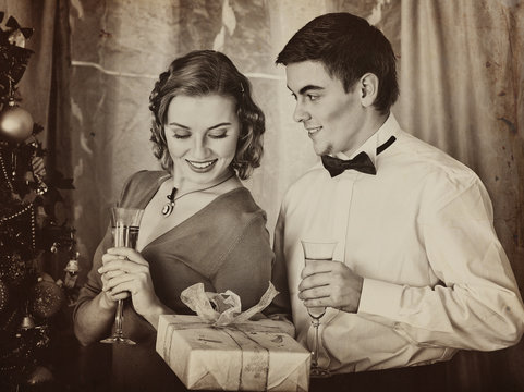 Old photo on yellow paper. Black and white retro. Young people decoration Christmas tree. Girl drink champagne. Man give gift box.