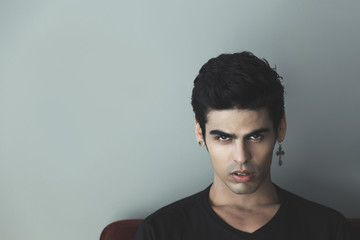 Portrait of attractive modern young white brazilian guy with black hair and cross-earring looking sullenly and saucily, gray wall behind