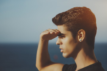 Portrait of young handsome brazilian guy with black hair and earring standing in front of ocean and...