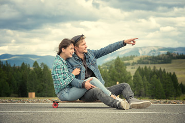 The guy with girl sit on a longboard. Man shows his hand forward. Road in the mountains.