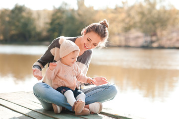 Smiling young mother sitting with baby girl 1 year old wearing casual knitted clothes outdoors. Motherhood.