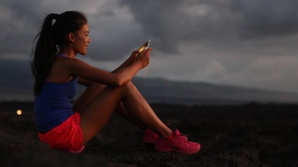 Woman using smart phone looking at fitness tracking app after running at night. Running sport and fitness runner woman relaxing outside looking at smartphone screen. Healthy lifestyle, Hawaii, USA. - Powered by Adobe