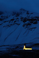 Naklejka na ściany i meble Vik Church in Iceland. Night photo with light church and dark blue snowy mountain. Winter scene from cold Iceland. Wild nature with building. Beautiful winter landscape.