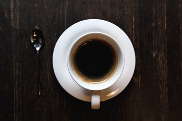 A dark, soft-focus image of a white coffee cup with black coffee and a tea spoon on a dark wooden table