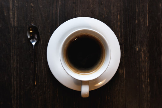 A dark, soft-focus image of a white coffee cup with black coffee and a tea spoon on a dark wooden table