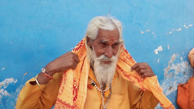 Indian sadhu saffron holy clothes putting on a turban against a blue wall 