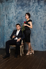 Fototapeta na wymiar a man in a blue suit with a wine glass sits on a chair brunette woman in a black dress with a rose standing next