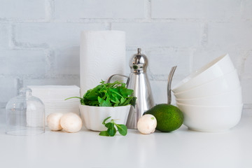 Cooking concept, kitchen interior - mushrooms. salad, green herbs, avocado, oil jug and dishes on white against white brick wall. Copy space.