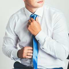 close-up of a man in a white shirt and blue tie standing near a white wall.young man adjusts his tie.