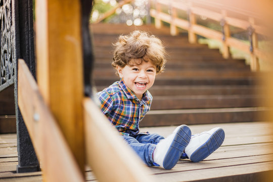 Beautiful sunshine toddler boy sitting on old wooden stairs and laughing joyfully while looking at camera. Portrait of adorable happy baby playing alone outside on warm summer day. Horizontal image.