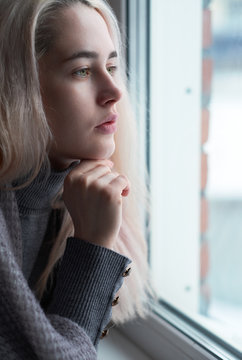 Young sad blonde woman with green pistachio colour eyes and without makeup is looking out of window