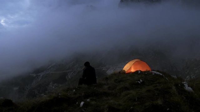 Wide shot of a man sitting next to his tent on a mountain top in the evening.
