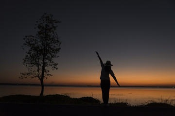 Silhouette of a beautiful woman wearing a hat spreading hand near big tree and reservoir, against sunset background.