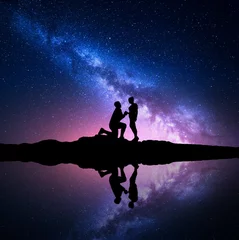 Poster Milky Way. Silhouettes of a man making marriage proposal to his girlfriend on the hill near the lake with starry sky reflection in water. Night landscape. Silhouette of couple. Milky way with people © den-belitsky