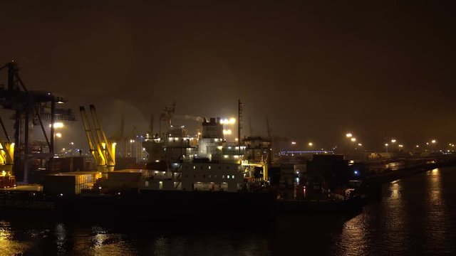 Ship leaving harbor sailing along the jetty pier with many crane spreaders and containers at the night
