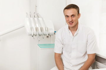 Dentist sitting on a chair near dental equipment and looking at the camera, which is in the workplace, in the dental clinic