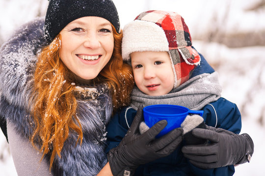 Close up portrait of baby sitting with young beautiful mother outside at snowy trees winter background and drinking hot tee. Happy family enjoying beautiful frosty days on picnic Horizontal photo.