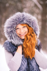 Portrait of beautiful young girl dressed in fur coat at winter trees background. Beauty woman having fun outside on snowy day. Close up of female smiling face with gorgeous long ginger hair.