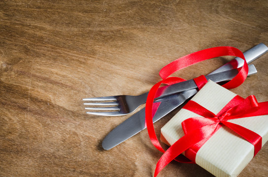 Present and Cutlery Decorated with Red Ribbon on Wooden Background.