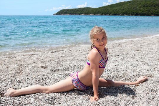 Cute Caucasian girl in swimsuit doing side split exercises on sea beach