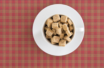 Top view of white coffee or tea cup full brown sugar cubes on red squared background. Diet unhealty sweet addiction concept