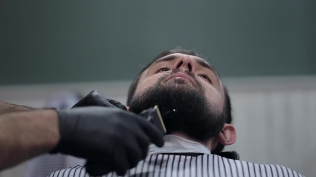 Male barber cutting beard with electric razor at a barber shop.