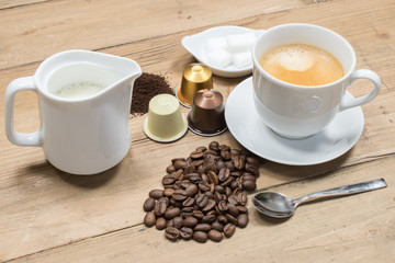 Coffee concept: Creamy cup of coffee, coffee beans, jug of milk, machine capsules and sugar cubes on wooden background.