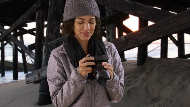 Portrait of young hipster woman holding camera, standing on beach by pier.