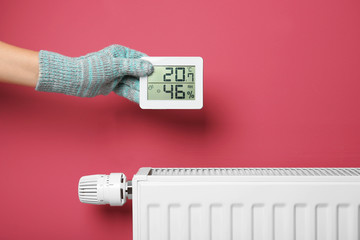Female hand in warm glove holding electronic thermometer near heating battery