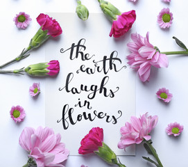 Inscription "THE EARTH LAUGHS IN FLOWERS" written on paper with flowers on white background