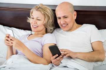 Mature boyfriend and girlfriend lying in bed socialising with mo