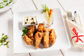 Grilled wings with vegetables and cutlery. Restaurant serving of fried chicken with creamy sauce on white plate, with fork and knife, wooden table. Junk food concept