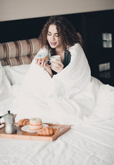 Smiling young girl 20-24 year old sitting wrapped in blanket having breakfast in bed with donuts...