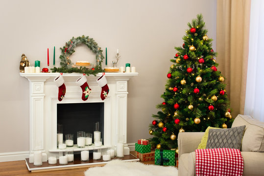 A new year decorated house with a fine tree full of colorful toys and a fireplace with candles and traditional socks hanging on it. Holiday decorated room with Christmas tree with presents under it.