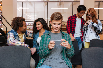 Attractive young man using smartphone while students standing and talking