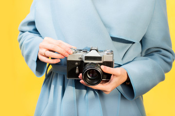 Woman in the blue coat holding retro camera on the yellow background. Close-up view with no face focused on the camera