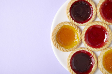 Overhead view of a plate full of assorted fruit jam tarts on a pastel purple background with blank space at side