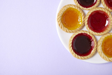 A plate full of assorted fruit jam tarts on a pastel purple background with blank space at side