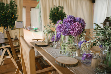 White and violet peonies stand in glass bottles on wooden dinner