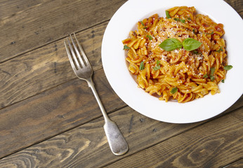 A dinner dish full of tomato and basil fusilli pasta on a rustic wooden dining table background with fork and blank space at side