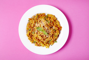 Aerial view of a dinner dish full of tomato and basil fusilli pasta on a bright pink background
